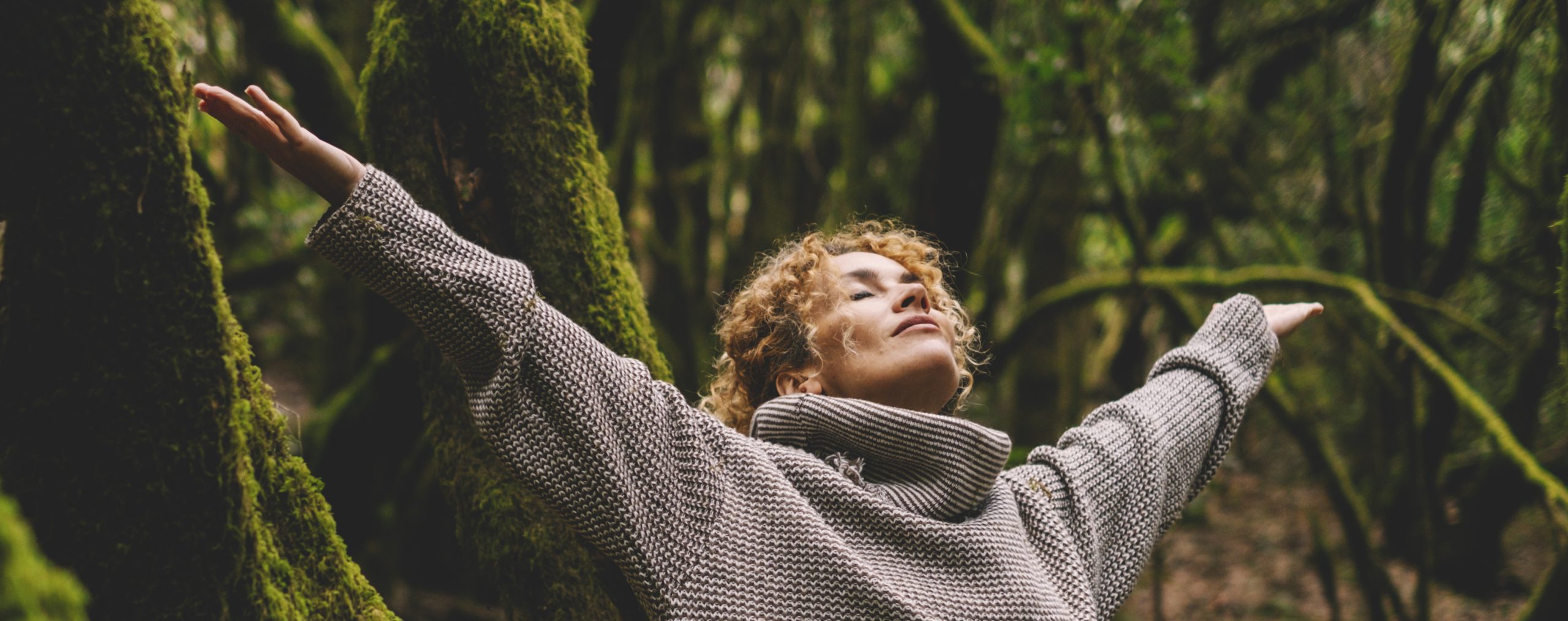 Celebrating freedom and nature love. One woman outstretching arms in the forest with green trees trunk and musk in background. Happiness and wellbeing in natural healthy lifestyle people in leisure